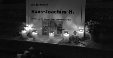 Das Bild zeigt eine schlichte schwarz-weiße Tafel in Erinnerung an Hans-Joachim Heidelberg. Vor der Tafel wurden Blumen abgelegt. Es wurden Kerzen vor der Tafel abgestellt und angezündet.