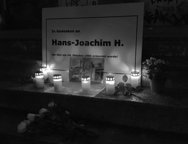 Das Bild zeigt eine schlichte schwarz-weiße Tafel in Erinnerung an Hans-Joachim Heidelberg. Vor der Tafel wurden Blumen abgelegt. Es wurden Kerzen vor der Tafel abgestellt und angezündet.