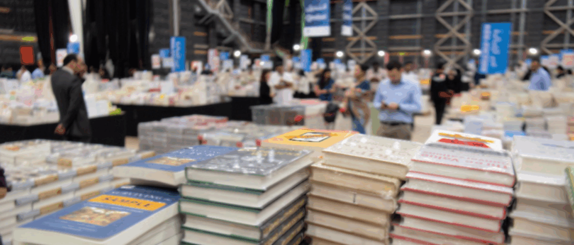 Zu sehen ist eine Messehalle mit vielen Besucher*innen und stapelweise Bücher.