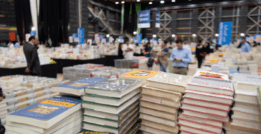 Zu sehen ist eine Messehalle mit vielen Besucher*innen und stapelweise Bücher.