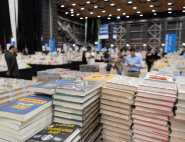 Zu sehen ist eine Messehalle mit vielen Besucher*innen und stapelweise Bücher.